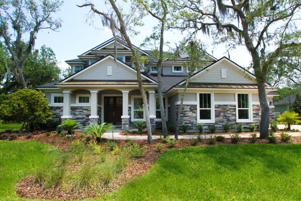 Elegant two-story custom home by Cornerstone Homes, showcasing a blend of stone and stucco finishes, gable roofs, and columned porch, nestled among mature trees with well-manicured landscaping.
